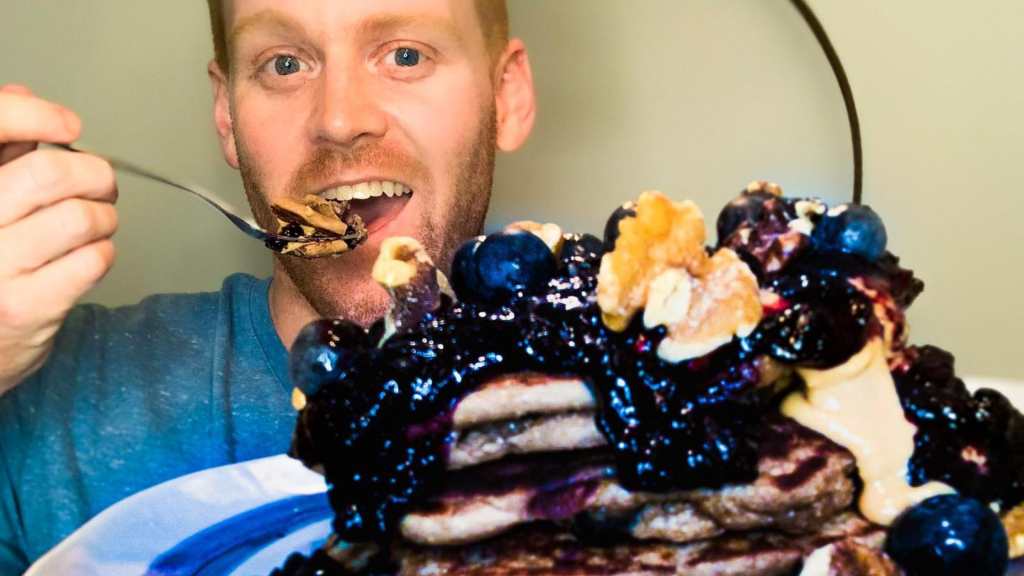 Man with a fork taking a bite of a large blueberry cream cake held in front of him, smiling at the camera.
